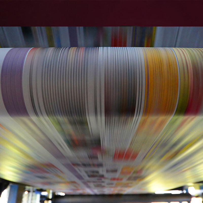 Nahaufnahme einer schnell laufenden Papierbahn in einer Rotationsdruckmaschine. Die bunten Druckmotive sind durch die hohe Geschwindigkeit als vertikale Streifen unscharf dargestellt